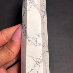 May include: A white Howlite crystal tower with gray and gold veining. The polished stone has a hexagonal shape and a pointed top. The crystal is held against a dark background.