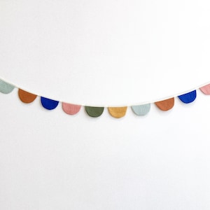 May include: A colorful garland featuring semi-circular fabric pieces in various colors, including blue, pink, and brown. The garland is strung on a white cord and is set against a plain white background. This decorative item is suitable for parties or room decor.