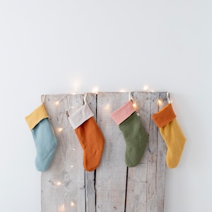 Op de afbeelding: Vier kleurrijke kerstsokken met contrasterende manchetten hangen aan een rustieke houten plank. De sokken zijn in blauw, oranje, groen en geel. Een lichtslinger voegt een feestelijke sfeer toe.