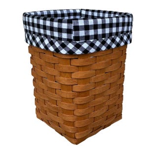 May include: A rectangular woven basket in a brown hue. The top edge is trimmed with a black and white gingham fabric. The basket is empty and set against a white backdrop.