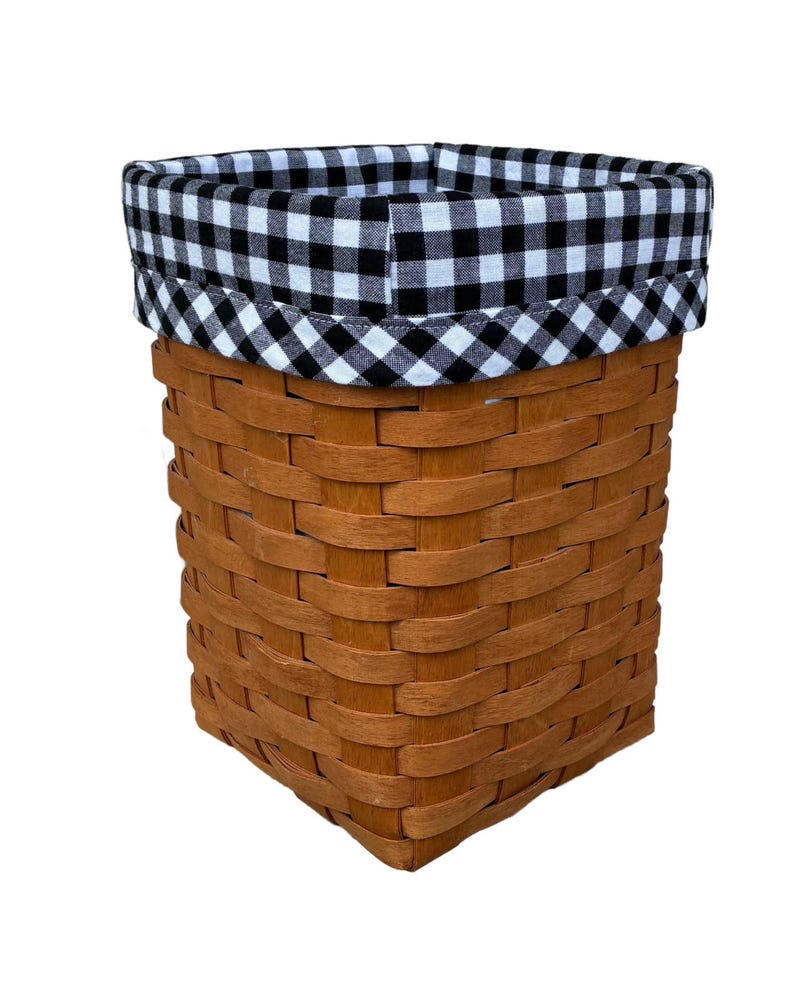 May include: A rectangular woven basket in a brown hue. The top edge is trimmed with a black and white gingham fabric. The basket is empty and set against a white backdrop.