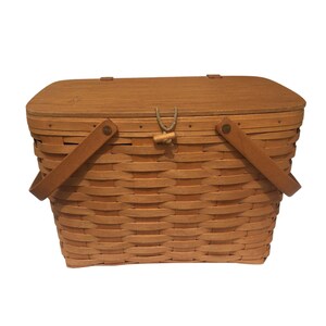 May include: A light brown woven picnic basket with a wooden lid and two attached handles. The lid is secured with a wooden toggle and a loop of rope. The basket is made of woven strips of wood.