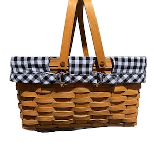 May include: A rectangular picnic basket with a woven wooden body and a black and white gingham fabric lining the top. It has a wooden handle and metal clasps. The basket is designed for carrying food and drinks.