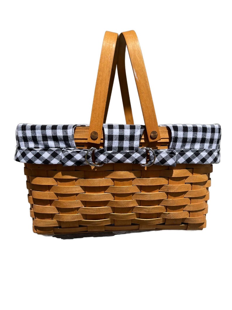 May include: A rectangular picnic basket with a woven wooden body and a black and white gingham fabric lining the top. It has a wooden handle and metal clasps. The basket is designed for carrying food and drinks.