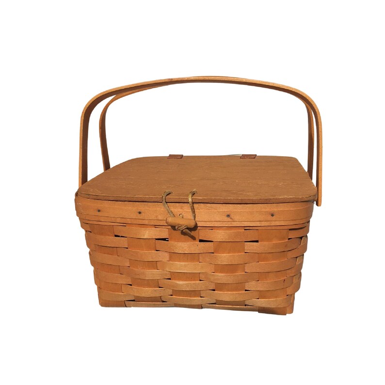 May include: A light brown, woven picnic basket with a wooden lid and a curved handle. The basket has a rectangular shape with rounded edges and a woven pattern. The lid is secured with a rope and loop closure.