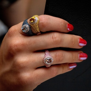 May include: Three rings with colorful gemstones set in a pave setting. The rings are on a hand with red nail polish.