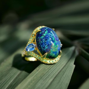 May include: A gold ring featuring a large, oval-shaped blue gemstone with green and blue inclusions. The ring is accented with smaller blue and green gemstones. The ring is set on a green leaf.