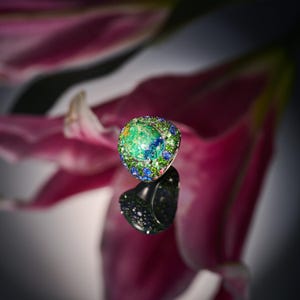 May include: A green and blue gemstone ring with a floral design. The ring is set in silver and features a large, round gemstone surrounded by smaller gemstones.