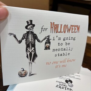 May include: A white greeting card featuring a skeleton wearing a top hat and coat, holding a lantern with a small pumpkin inside. Text reads: for HALLOWEEN i'm going to be mentally stable no one will know it's me. A small jack-o'-lantern sits at the feet.