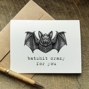 May include: A white greeting card with a detailed black and white illustration of a bat with its wings spread. The text "batshit crazy for you" is printed below the bat. A pen and envelope are in the background.