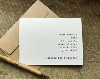 May include: A white greeting card with black typewriter-style text that reads: what does it mean if the holy water sizzles when it hits your skin? (asking for a friend). The card is paired with a brown envelope and a wooden pen.