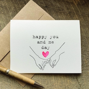 May include: A white greeting card with the text "happy you and me day" and a pink heart. The card features line art of two hands holding pinky fingers. A brown envelope and a pen are also in the image.