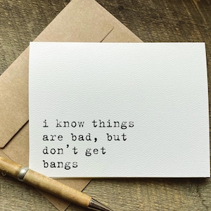 May include: A white greeting card with the text "I know things are bad, but don't get bangs" in black typewriter font. The card is on a brown envelope, with a wooden pen on a wooden surface.