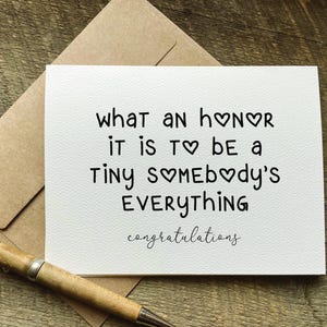 May include: A white greeting card with the text "what an honor it is to be a tiny somebody's everything congratulations" in black. A pen and envelope are next to the card. The card is on a wooden surface.