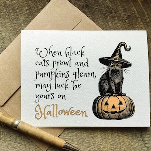 May include: A white greeting card featuring a black cat wearing a witch hat perched on a carved orange pumpkin. The text reads: When black cats prowl and pumpkins gleam, may luck be yours on Halloween. The card is paired with a brown envelope.