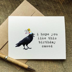 May include: A birthday card with a black crow wearing a party hat. The card reads "i hope you like this birthday cawed". A pen and envelope are in the background.
