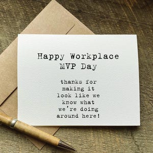 May include: A white card with the text "Happy Workplace MVP Day" and a message thanking someone for their work. The card is on a wooden surface with a pen and a brown envelope.