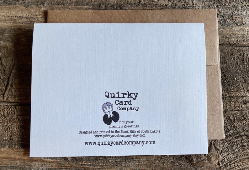 May include: A white card with black text that reads "Quirky Card Company" and a cartoon image of a woman with a surprised expression. The text also reads "not your granny's greetings" and "Designed and printed in the Black Hills of South Dakota" and includes the website address "www.quirkycardcompany.com".