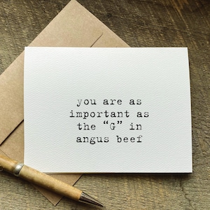 May include: A white greeting card with black typewriter-style text that reads, you are as important as the G in angus beef. The card is paired with a brown kraft paper envelope and a wooden pen, all resting on a rustic wooden surface.