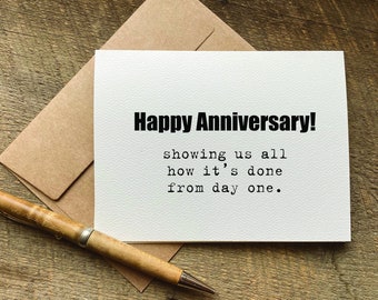 May include: A white card with the text "Happy Anniversary! showing us all how it's done from day one." on a brown wooden surface with a brown envelope and a wooden pen.