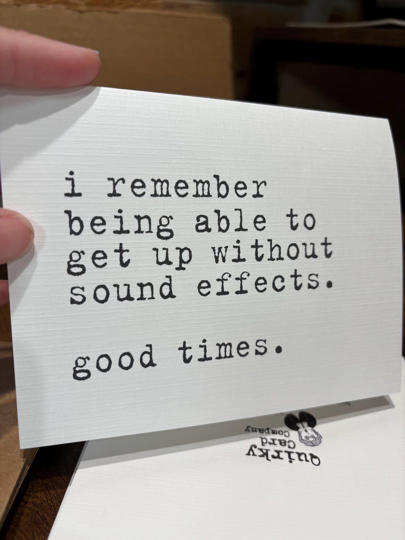 May include: A white rectangular greeting card with a textured surface featuring black typewriter-style text that reads: i remember being able to get up without sound effects. good times. The card has a clean, minimalist design with black lettering.