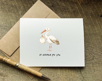 Puede incluir: Una tarjeta de felicitación blanca con una ilustración en acuarela de una cigüeña blanca con pico naranja que lleva un pequeño hatillo. Debajo de la cigüeña, el texto "so storked for you" aparece en letras negras sin remates.
