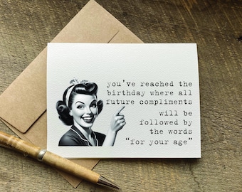 May include: A greeting card with a vintage illustration of a woman pointing, accompanied by text that reads, "you've reached the birthday where all future compliments will be followed by the words 'for your age.'" The card is on a wooden surface.