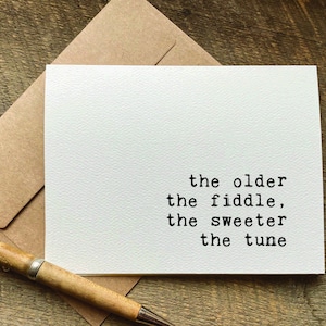 May include: A white card with black text that reads "the older the fiddle, the sweeter the tune". The card is sitting on a brown envelope.