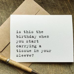 May include: A white greeting card with the text "is this the birthday when you start carrying a tissue in your sleeve?" is displayed on a wooden surface. A brown envelope and a pen are also present.