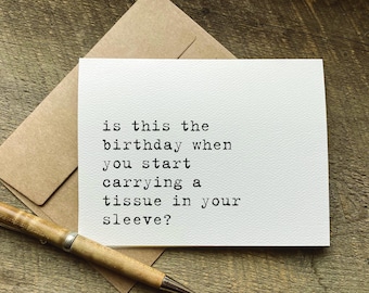 Può includere: Un biglietto di auguri bianco con il testo "è questo il compleanno in cui inizi a portare un fazzoletto nella manica?" è esposto su una superficie di legno. Sono presenti anche una busta marrone e una penna.