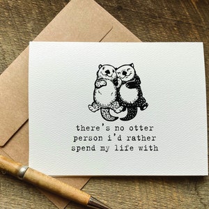 May include: A white card with a black and white illustration of two otters holding each other. The text on the card reads "there's no otter person I'd rather spend my life with."