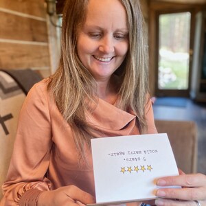 May include: A woman smiles while holding a card that says "5 stars. Would make a great gift." with five gold stars.