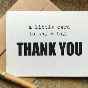 May include: A white card with the text "a little card to say a big THANK YOU" in black. The card is on a brown envelope, with a wooden pen in the foreground. The card has a textured surface.