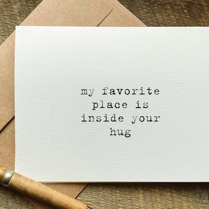 May include: A white rectangular greeting card with a textured surface featuring the black typewriter-style text "my favorite place is inside your hug" centered on the front. The card is paired with a brown envelope and a wooden pen.