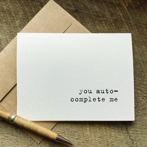 May include: A white greeting card with the text "you auto-complete me" in a typewriter font. The card is on top of a brown envelope, with a wooden pen in the foreground.