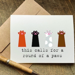 May include: A greeting card with a white background and four paw prints in different colors. The paw prints are orange, black, gray with white spots, and brown. The text on the card reads "this calls for a round of a paws".