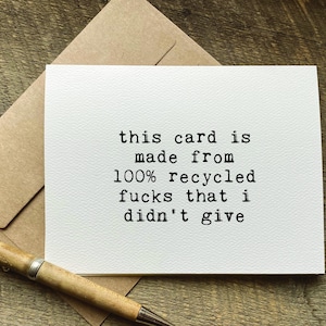 May include: A white greeting card with the text "this card is made from 100% recycled fucks that i didn't give" in black typewriter font. A brown envelope and a pen are also in the image.