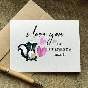 May include: A greeting card with a watercolor skunk illustration holding a heart, accompanied by the phrase "I love you so stinking much." Pink and tan hearts accent the design. A pen and envelope are also visible.
