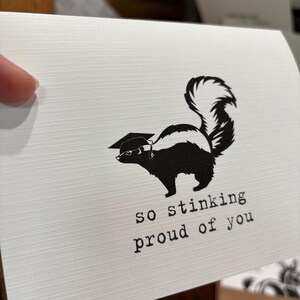 May include: A white greeting card with a textured surface featuring a black graphic of a skunk wearing a graduation cap. Below the skunk, the text reads "so stinking proud of you" in a typewriter-style font. The card is held against a blurred background.