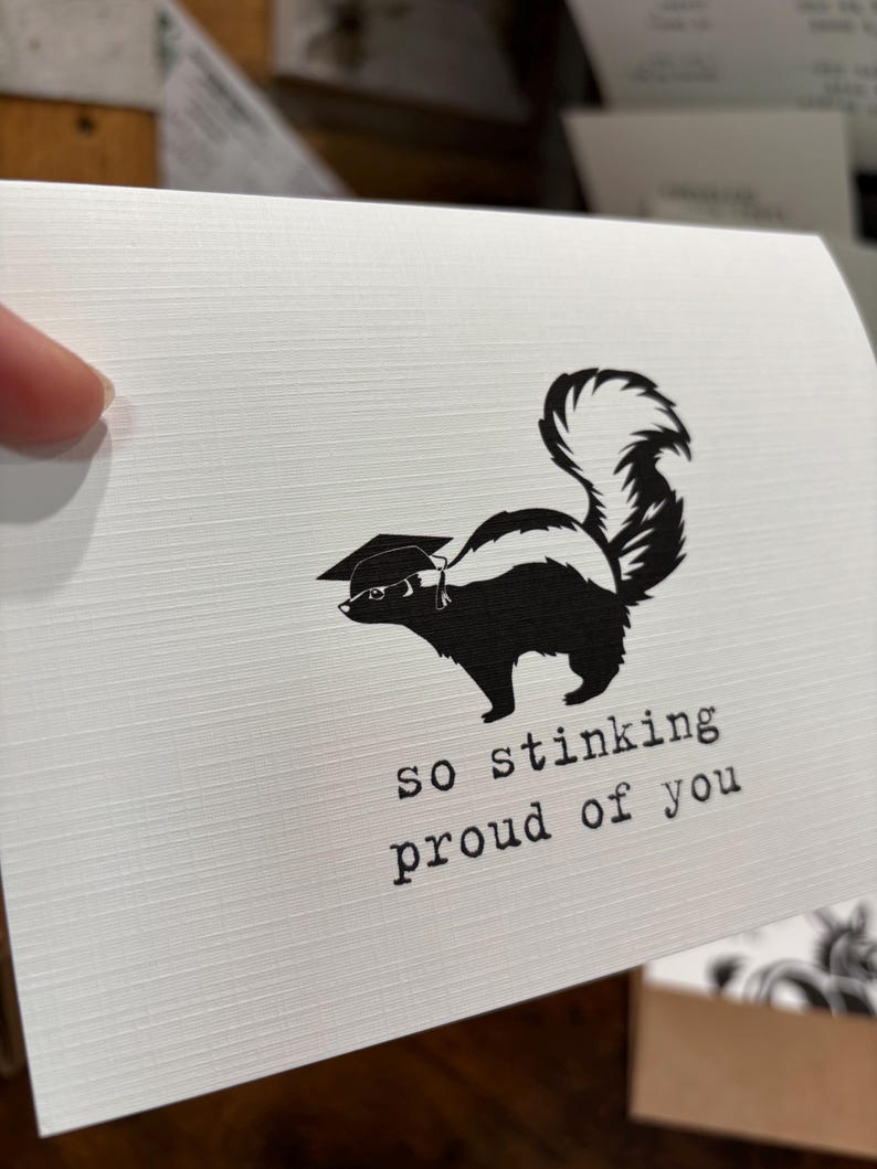 May include: A white greeting card with a textured surface featuring a black graphic of a skunk wearing a graduation cap. Below the skunk, the text reads "so stinking proud of you" in a typewriter-style font. The card is held against a blurred background.