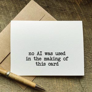 May include: A white greeting card with the text "no AI was used in the making of this card" in black. The card is on a brown envelope, with a wooden pen in the foreground.