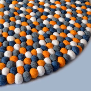 May include: A round, colourful rug made of felt balls. The rug is made up of orange, white, grey, and blue felt balls arranged in a repeating pattern.