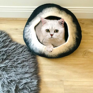 May include: A white cat with yellow eyes sits inside a black and white felt cat bed. The bed is round and has a hole in the center. The cat is looking directly at the camera.