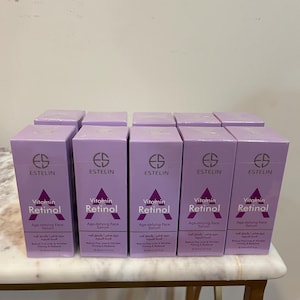 Puede incluir: Varias cajas de suero facial antiedad Estelin Vitamin Retinol están dispuestas sobre una superficie. Las cajas son de color lila claro con texto blanco y un logotipo. El texto incluye las palabras "Vitamin Retinol" y "Age-defying Face Serum".
