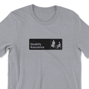 May include: A heather grey t-shirt featuring a black and white graphic of a sign that reads "Quality Assurance" with two stick figures, one standing and one sitting at a desk.