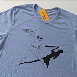 May include: A light blue t-shirt with a black graphic of a person playing tennis with their shadow behind them. The text "rhinotaur" is printed on a yellow tag attached to the shirt.