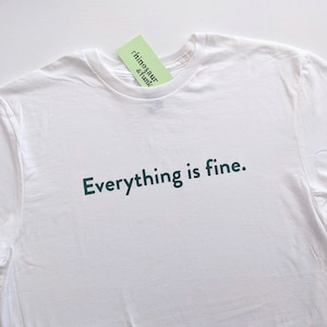 May include: White t-shirt with the text "Everything is fine." printed in dark green on the front. The shirt has a tag attached to the collar that reads "rhinosaure & funk".