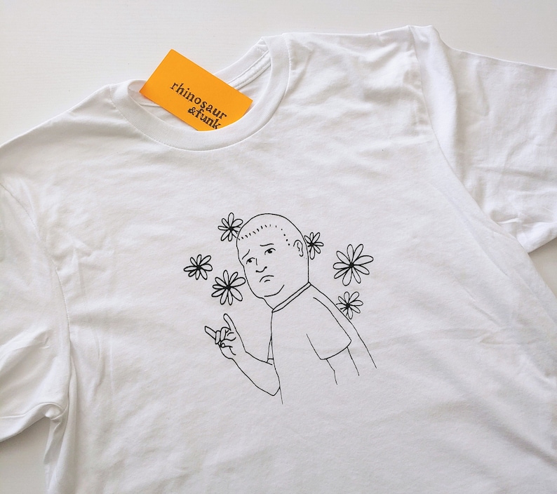 May include: White t-shirt with a black line drawing of a cartoon character with a sad expression and flowers around the head. The text "rhinosaur & funk" is on a yellow tag.