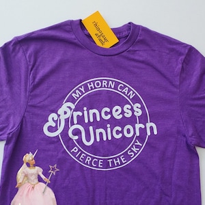 May include: Purple t-shirt with a white circle graphic that says "My horn can pierce the sky" and "Princess Unicorn" in a stylized font. A Barbie doll wearing a pink dress and holding a wand is visible in the bottom left corner.