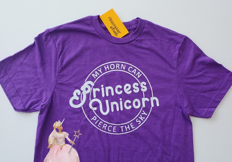 May include: Purple t-shirt with a white circle graphic that says "My horn can pierce the sky" and "Princess Unicorn" in a stylised font. A Barbie doll wearing a pink dress and holding a wand is visible in the bottom left corner.